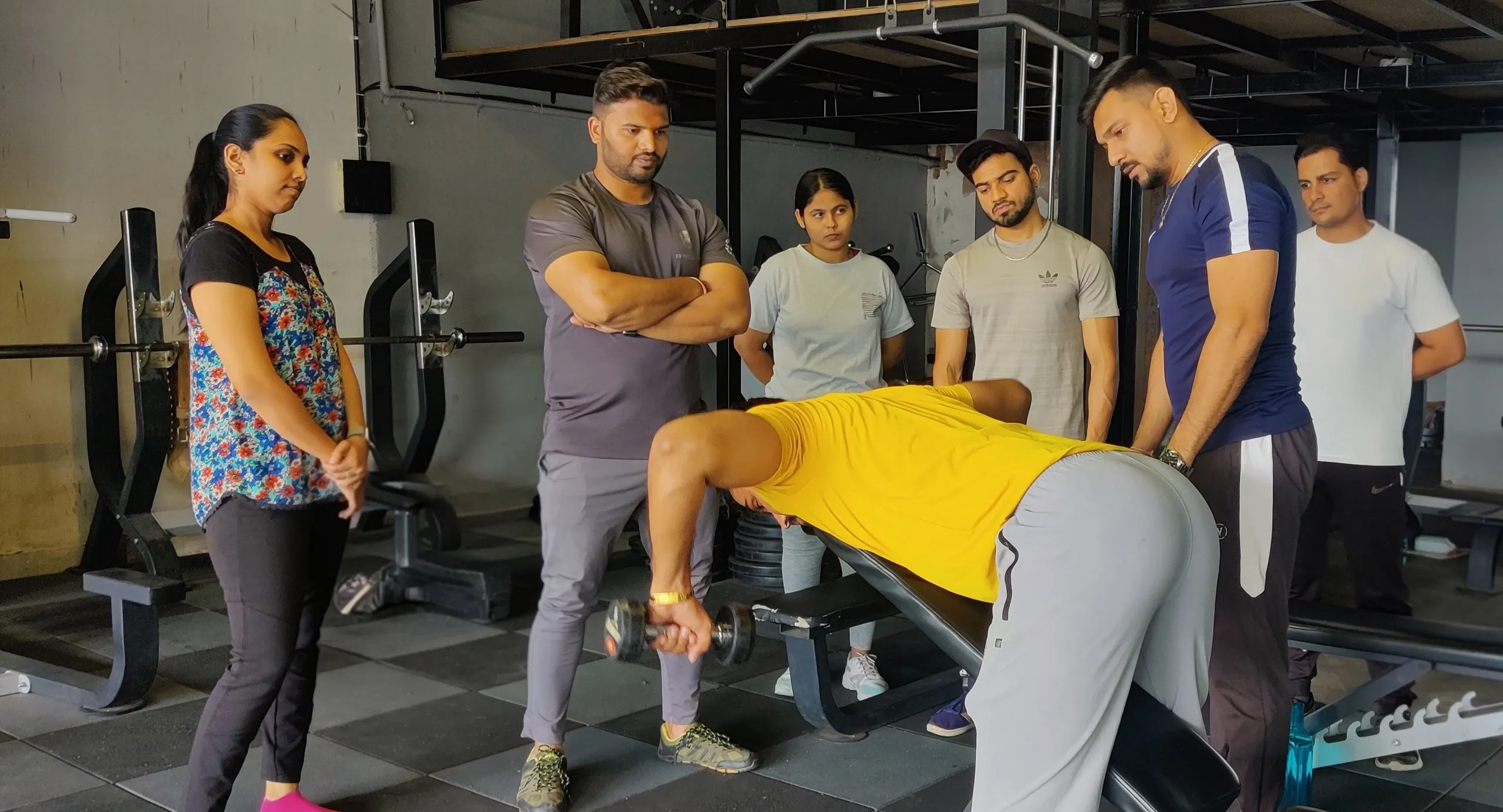 IHFE instructor correcting a trainee's dumbbell row form on the gym floor while student personal trainers watch